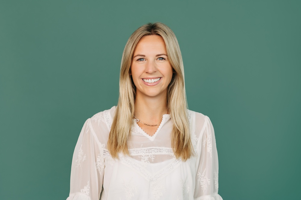 Eine Frau vor einem grünem Hintergrund. Sie trägt eine weiße Bluse. Sie hat blonde glatte lange Haare und lächelt freundlich